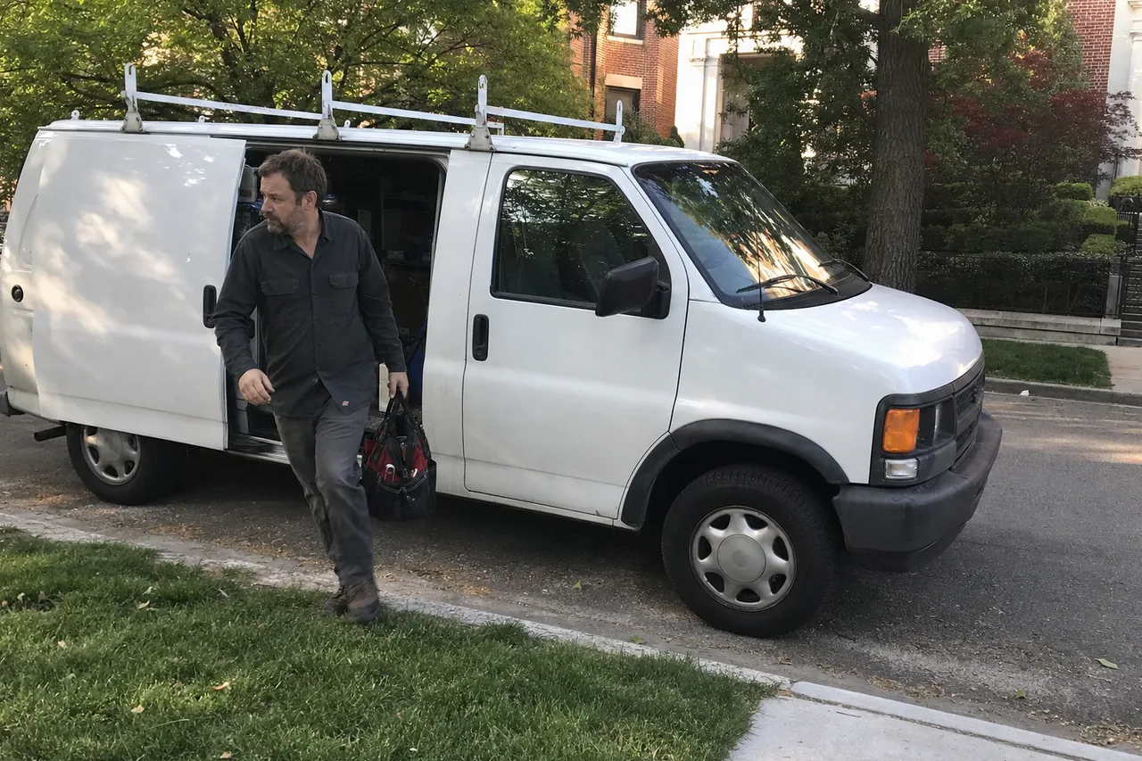A service van parked under mature street trees while a technician unloads his tool bag